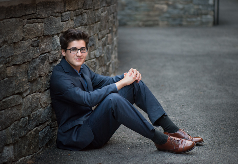 photo of a high school senior guy in a suit sitting by the stone wall, taken by Marika H Photography in Williamsburg, VA