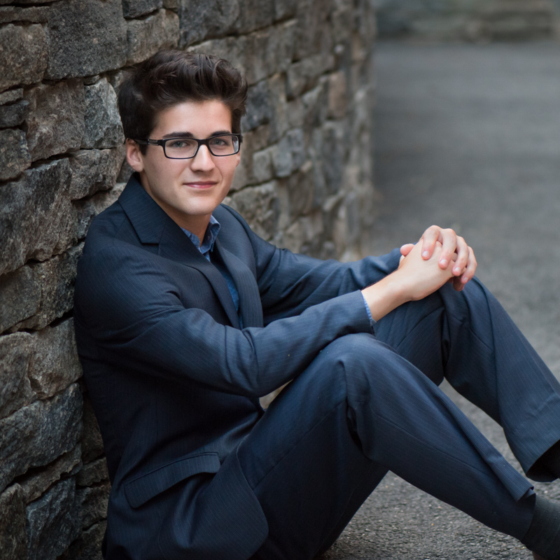 photo of a high school senior guy in a suit sitting by the stone wall, taken by Marika H Photography in Williamsburg, VA