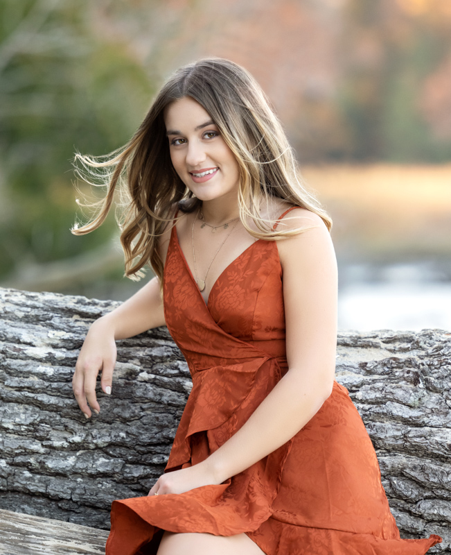 A photo of a high school senior girl sign on the log by the water in York River State Park, wearing a copper color dress, taken by Marika H Photography in Williamsburg, VA